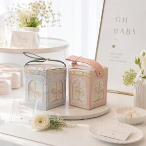 May include: Two decorative boxes, one blue and one pink, with carousel horse designs, sit on a marble surface. A sign reads "OH BABY" and "WELCOME TO OUR BABY SHOWER." The scene includes flowers and a "Thank You" card, suggesting a baby shower theme.