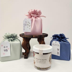Puede incluir: Tres bolsas de tela con cordón en rosa pastel, verde salvia y azul marino, cada una con una etiqueta blanca con texto en coreano. Un frasco de plástico transparente etiquetado Fruit & Nuts se sitúa delante sobre un pedestal de madera.