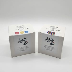 May include: Two white square boxes with Korean text and colorful accents. The boxes have the text "THE MAUM" at the top and the Korean word "첫출" in large black letters. The boxes are on a white background.