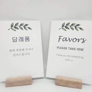 Puede incluir: Dos tarjetas blancas con texto y soportes de madera. Una tarjeta dice "Favors" con "Please Take One" y "Thank you for celebrating with us." La otra tarjeta tiene texto coreano y una ramita de hojas.