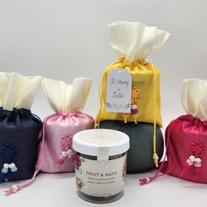 May include: Four small gift bags in yellow, pink, blue, and red satin with white fabric tops. Each bag has a decorative knot and a tag. A glass jar of fruit and nuts sits in front of the bags.