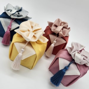 May include: Four square fabric gift wraps in navy, yellow, rust, and mauve, each featuring a contrasting cream-colored top knot and a decorative hanging tassel. The fabric is folded into a structured box shape with a smooth, satin-like finish.
