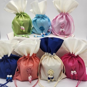 May include: A collection of seven colorful drawstring pouches. Each pouch is a different color, including green, blue, pink, coral, cream, and fuchsia. They feature a white top and decorative knot accents with tassels.