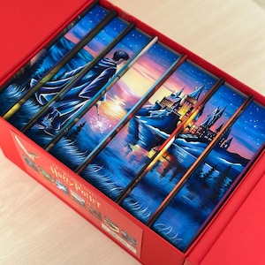 May include: A red box set containing seven books with spines that form a continuous illustration of a night landscape, a figure in a robe holding a wand, and a castle. The text J.K. Rowling Harry Potter is printed on the side of the red box.