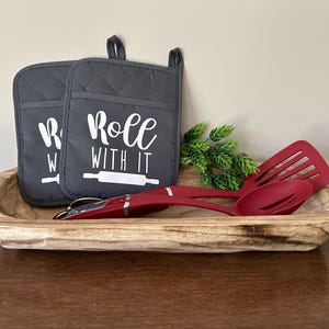 May include: Two gray oven mitts with white text that reads "Roll with it" and a white rolling pin graphic. The mitts are in a wooden bowl with two red kitchen utensils.