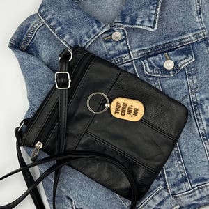 May include: Black leather crossbody bag with a silver-tone zipper and strap, featuring a wooden keychain that reads "THAT CURB HIT ME." The bag rests on a blue denim jacket.