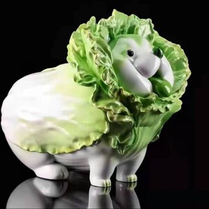 以下が含まれることがあります： 緑色のレタスの頭を持つ犬のような生き物の気まぐれなセラミック彫刻。体は白く、レタスは緑と黄色の色合いです。彫刻は光沢のある仕上がりです。