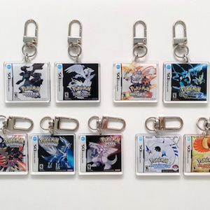 Op de afbeelding: Een set van tien sleutelhangers met verschillende Pokémon-spelhoezen. De sleutelhangers zijn zilverkleurig met doorzichtige plastic hoezen. De hoezen tonen de Pokémon-spellen Diamant, Parel, Platina, Zwart, Wit, Zwart 2, Wit 2, HeartGold, SoulSilver en Omega Ruby.