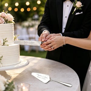 Puede incluir: Un cuchillo para pastel de plata y una pala para pastel a juego, ambos grabados con el texto "Angelica & James September 08, 2023". El cuchillo se utiliza para cortar un pastel de bodas blanco de dos niveles decorado con rosas rosas y blancas.
