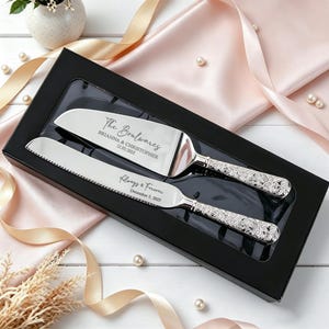 May include: A silver cake knife and server set with ornate floral handles, presented in a black box. The server is engraved with "The Boulwares, Brianna & Christopher, 12.03.2025" and the knife is engraved with "Always & Forever, December 3, 2025."