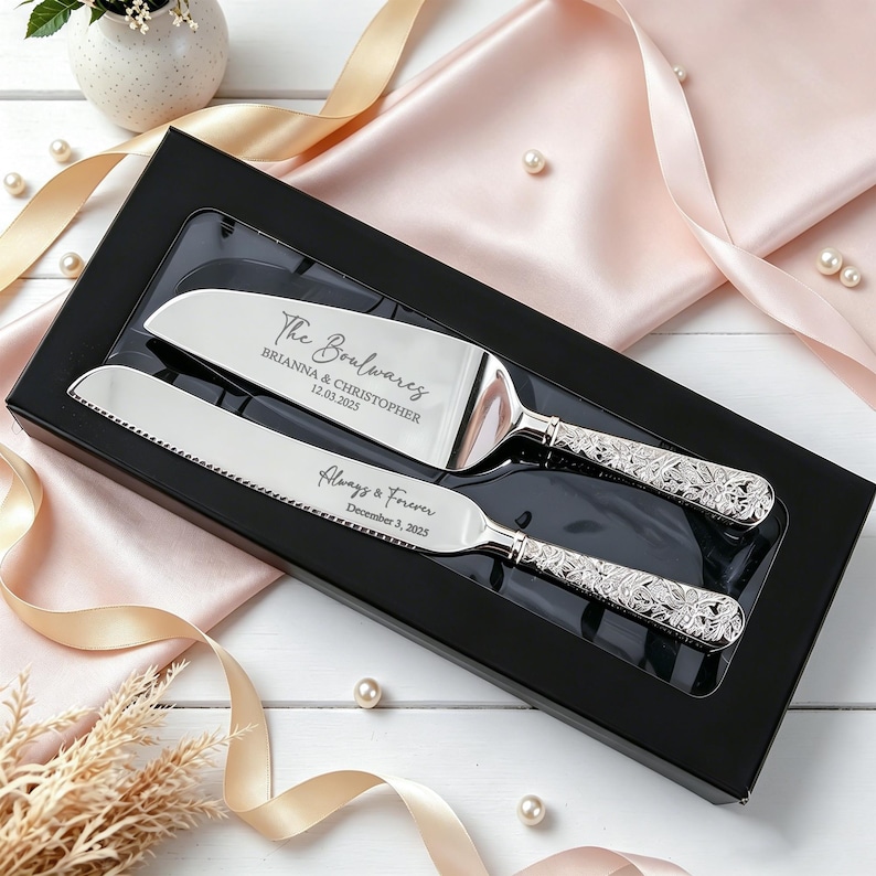 May include: A silver cake knife and server set with ornate floral handles, presented in a black box. The server is engraved with "The Boulwares, Brianna & Christopher, 12.03.2025" and the knife is engraved with "Always & Forever, December 3, 2025."