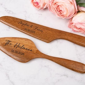 Könnte beinhalten: Ein Tortenmesser- und Tortenheber-Set aus Holz mit glatter, polierter Oberfläche. Das Messer ist mit dem Text Angelica & James 06.06.2025 graviert, und der Heber trägt die Gravur The Heilmann 06.06.2025 in einer dunklen Schreibschrift.