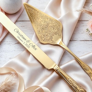 May include: A gold wedding cake knife and server set. The knife is engraved with "Christina & Alex" and "November 1 2025". The server has an ornate floral design. The set rests on a satin cloth with roses.