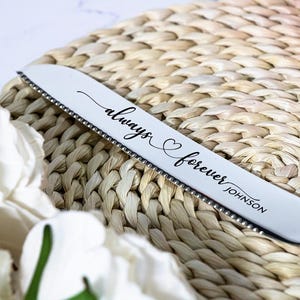 May include: A silver-toned serrated cake knife featuring the engraved script text always forever JOHNSON with a small heart symbol between the words. The blade has a metallic finish and a distinct serrated edge along the bottom side.