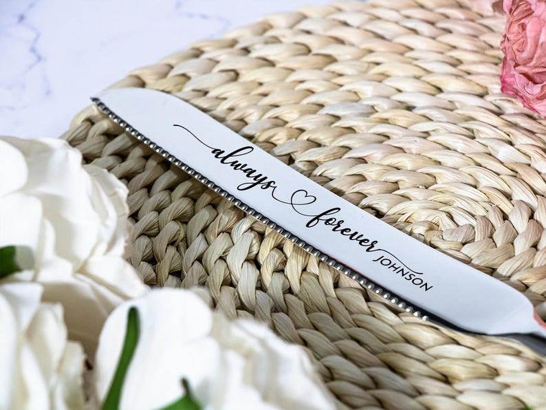 May include: A silver-toned serrated cake knife featuring the engraved script text always forever JOHNSON with a small heart symbol between the words. The blade has a metallic finish and a distinct serrated edge along the bottom side.