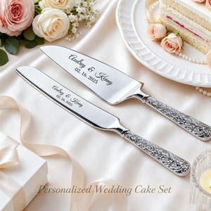 Pode incluir: Um conjunto de faca e espátula para bolo em prata com cabos de design floral ornamentado. As lâminas estão gravadas com o texto Audrey & Kenny e a data 02.16.2025 em fonte cursiva preta. As superfícies metálicas são polidas e muito reflexivas.
