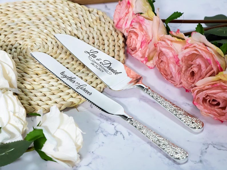 May include: A silver cake knife and server set with ornate, textured handles. The knife is engraved with the text together forever, and the server is engraved with the names Lea & Daniel and the date 06.24.2024 in a cursive script font.