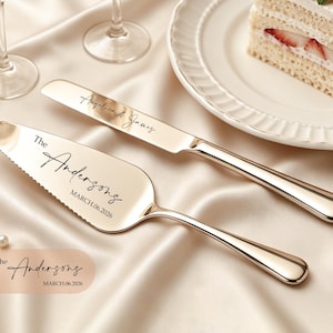 Puede incluir: Un juego de cuchillo y pala para pastel en tono dorado con grabado personalizado. El cuchillo lleva los nombres Angelica & James, mientras que la pala muestra el texto The Andersons y la fecha MARCH.06.2026 en una fuente cursiva negra.