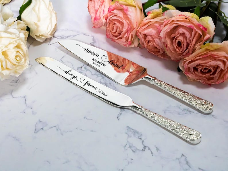 May include: A silver cake knife and server set with textured handles. The knife is engraved with "always forever JOHNSON" and the server features the names "Monica & Daniel", the surname "JOHNSON", and the date "04 15 22" in black script lettering.