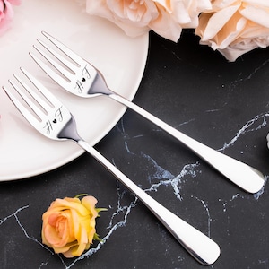 Puede incluir: Dos tenedores plateados con las iniciales "H.F" y un símbolo de corazón grabados, colocados sobre un plato blanco. La imagen presenta arreglos florales con rosas rosas y melocotón, sobre un fondo de mármol negro. Un regalo ideal para bodas.