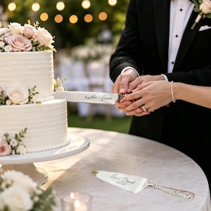 May include: A silver cake knife engraved with the text together forever and a heart symbol, and a matching silver cake server engraved with the text Lea & Daniel 06.24.2024, both featuring ornate handles and polished metallic surfaces.