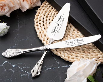 May include: Silver cake knife and server set with ornate handles, personalized with the names "Nadine & Vance" and the date "October 25, 2025". The set rests on a woven mat, with white and peach roses nearby.