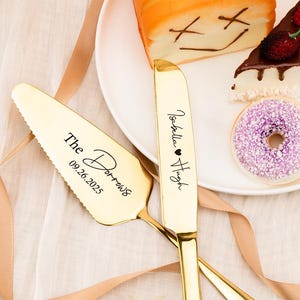 May include: Gold-colored wedding cake server and knife set. The server is engraved with "The Darrows" and the date "09.26.2025". The knife is engraved with "Isabella ❤ Hugh". The set is displayed with cake and a donut.