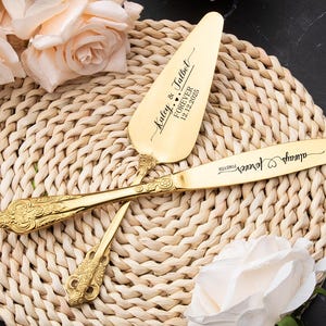 以下が含まれることがあります： 「Kaley & Talbot FOREVER 12.12.2025」と「always forever」の文字が刻まれた、金色の結婚式のケーキナイフとサーバーのセット。装飾的なハンドルが付いています。織りのプレースマットの上に置かれ、花に囲まれています。