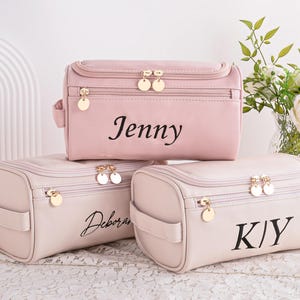May include: Three personalised cosmetic bags in pink and cream. The bags have gold-tone zips and handles. The top bag is pink and says "Jenny". The other two bags are cream-coloured and say "Deborah" and "KIY".