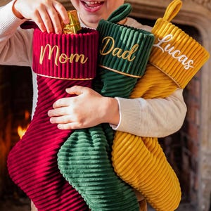 Può includere: Tre calze natalizie in velluto a coste nei colori rosso, verde scuro e giallo senape. Ogni calza presenta un risvolto con bordo dorato e nomi ricamati: Mom in oro, Dad in oro e Lucas in corsivo bianco sulle rispettive calze natalizie.
