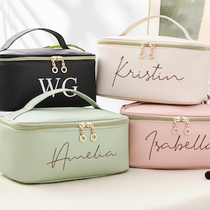 May include: Four personalized cosmetic bags in black, cream, mint green, and pink. Each bag has a gold zipper and handle. Names are scripted on the bags: WG, Kristin, Amelia, and Isabella. A makeup brush set is visible.