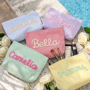 May include: Five rectangular seersucker pouches in light blue, purple, red, mint green, and yellow, each featuring a personalized name in raised fabric lettering. The pouches are arranged with makeup brushes, a tube, and a bottle nearby.