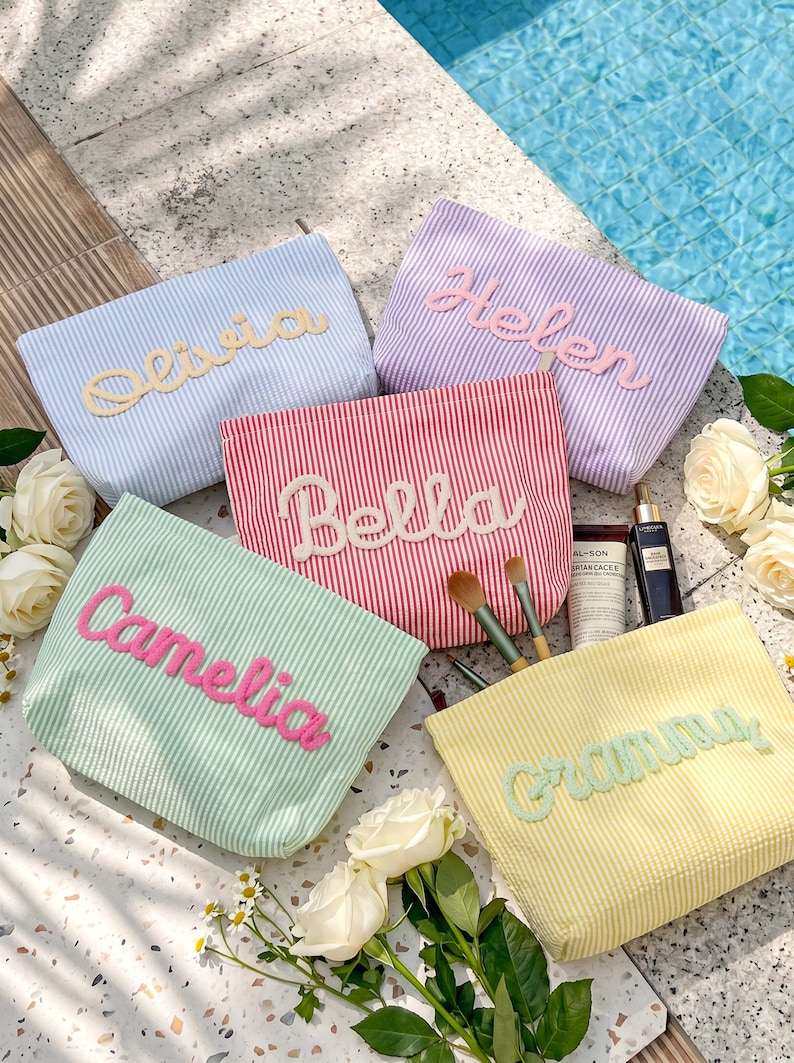 May include: Five rectangular seersucker pouches in light blue, purple, red, mint green, and yellow, each featuring a personalized name in raised fabric lettering. The pouches are arranged with makeup brushes, a tube, and a bottle nearby.