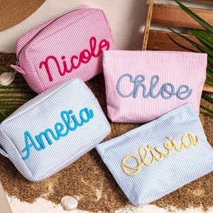May include: Four rectangular zippered pouches made of seersucker fabric with thin stripes. Two pouches are pink and white, and two are blue and white. Each pouch features a name embroidered in cursive script: Nicole, Chloe, Amelia, and Olivia.