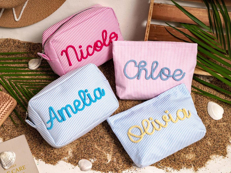 May include: Four rectangular zippered pouches made of seersucker fabric with thin stripes. Two pouches are pink and white, and two are blue and white. Each pouch features a name embroidered in cursive script: Nicole, Chloe, Amelia, and Olivia.