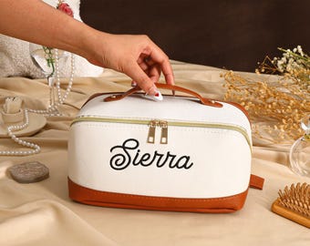May include: A white cosmetic bag with brown leather trim and a matching handle. The bag features the name "Sierra" in black script. The bag has a gold zipper and is sitting on a beige surface.