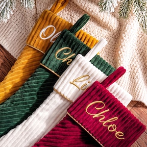 May include: Four corduroy Christmas stockings in mustard yellow, forest green, white, and deep red. Each stocking features a hanging loop and is embroidered with a name in cursive script: Oliver, Charlie, Liam, and Chloe, respectively.