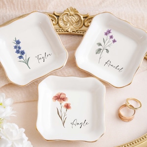 Op de afbeelding: Drie witte keramische ringschalen met gouden rand, elk met een bloemenontwerp en een naam: Taylor, Rachel en Angie. Er zijn ook twee gouden ringen aanwezig. De schalen zijn gerangschikt op een decoratief dienblad.
