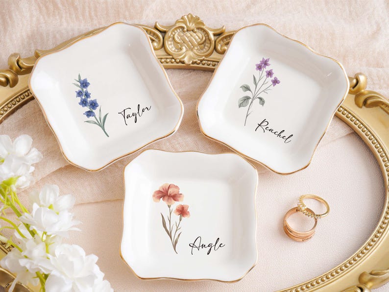 Op de afbeelding: Drie witte keramische ringschalen met gouden rand, elk met een bloemenontwerp en een naam: Taylor, Rachel en Angie. Er zijn ook twee gouden ringen aanwezig. De schalen zijn gerangschikt op een decoratief dienblad.