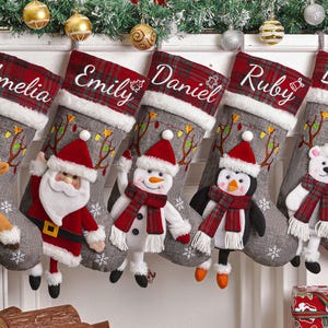 May include: Five Christmas stockings, each personalised with a name: Amelia, Emily, Daniel, Ruby, and Bella. The stockings have a grey body, red plaid cuff, and a plush character: a reindeer, Santa, a snowman, a penguin, and a polar bear.