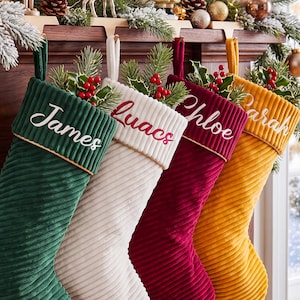 Pode incluir: Quatro meias de Natal de veludo cotelê nas cores verde-escuro, branco, vermelho profundo e amarelo-mostarda. Cada meia apresenta um nome bordado em letra cursiva branca ou vermelha no punho e está preenchida com ramos de pinheiro e bagas vermelhas.