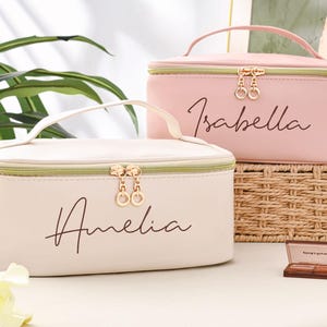 May include: Two personalized makeup bags with gold zippers. The top bag is pink and says "Isabella". The bottom bag is cream and says "Amelia".