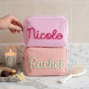 Puede incluir: Dos estuches rectangulares con cremallera hechos de tela a rayas. El estuche superior es rosa y blanco con el nombre Nicole en letras rosas en relieve. El estuche inferior es naranja y blanco con el nombre Rachel en letras verde claro.
