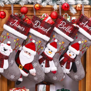 May include: Four grey fabric Christmas stockings with red and green plaid cuffs featuring the names James, Daniel, Bella, and Amelia. Each stocking displays a plush character: a polar bear, Santa Claus, a snowman, and a penguin, all wearing scarves.