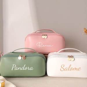 May include: Three cosmetic bags in pastel colors: pink, green, and white. Each bag has a top handle, a zipper closure with gold-tone pulls, and a name printed on the front. The pink bag says "Renee", the green bag says "Pandora", and the white bag says "Salome".