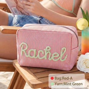 May include: A rectangular zippered pouch featuring a red and white pinstripe pattern with the name Rachel embroidered in mint green yarn across the front. The bag includes a black zipper closure and is labeled with Bag:Red-A and Yarn:Mint Green.