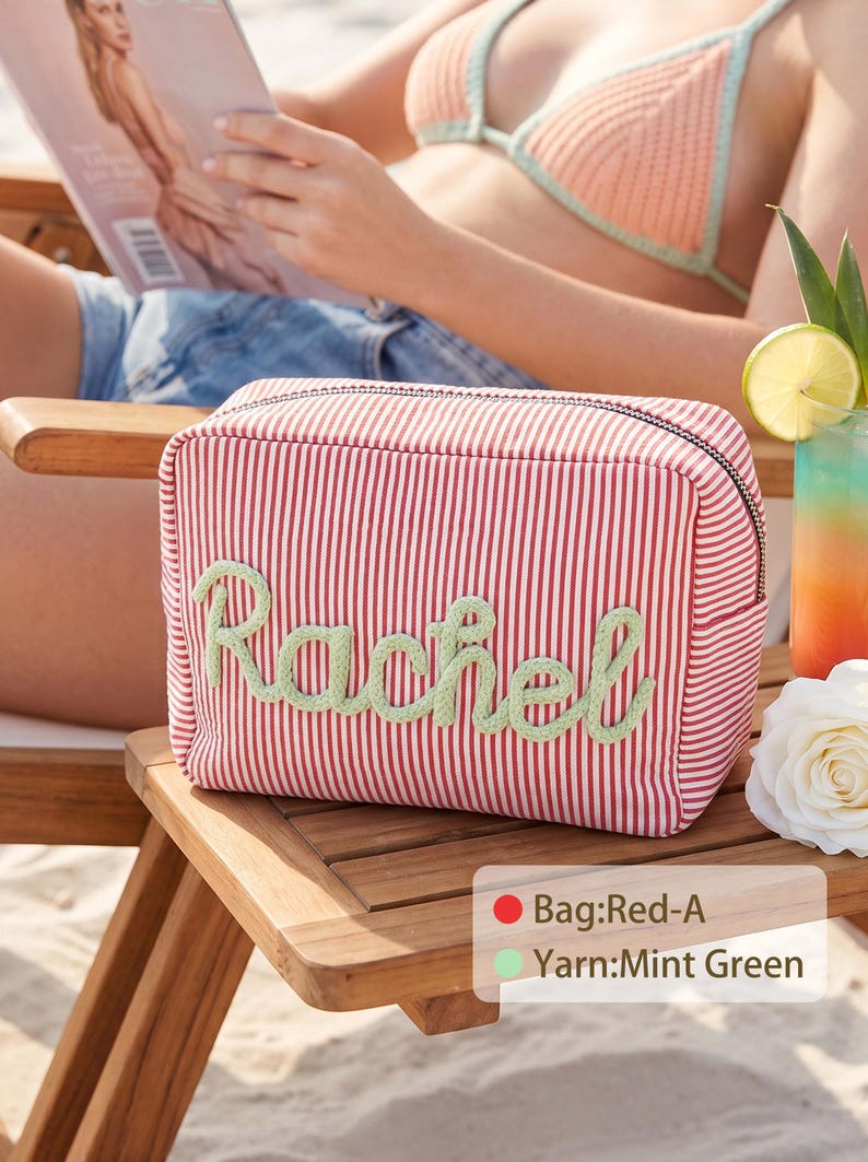 May include: A rectangular zippered pouch featuring a red and white pinstripe pattern with the name Rachel embroidered in mint green yarn across the front. The bag includes a black zipper closure and is labeled with Bag:Red-A and Yarn:Mint Green.