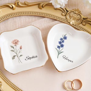 Op de afbeelding: Twee witte keramische ringenschaaltjes, een vierkante en een hartvormige, met gouden rand. Het vierkante schaaltje heeft een rozenontwerp en de naam "Sophia". Het hartvormige schaaltje heeft een blauw bloemenontwerp en de naam "Harper". Twee ringen op de voorgrond.