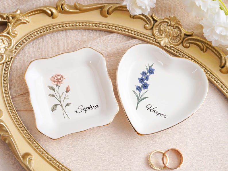 Op de afbeelding: Twee witte keramische ringenschaaltjes, een vierkante en een hartvormige, met gouden rand. Het vierkante schaaltje heeft een rozenontwerp en de naam "Sophia". Het hartvormige schaaltje heeft een blauw bloemenontwerp en de naam "Harper". Twee ringen op de voorgrond.