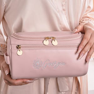 May include: A light pink cosmetic bag with gold-toned zippers and hardware. The bag features the name "Georgia" embroidered in a script font with a daisy design. The bag is held in front of a person wearing a light pink robe.